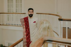A groom in white sherwani and red dupatta poses on an elegant staircase.