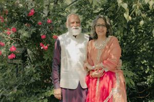 A couple in traditional Indian attire pose together in a garden with pink flowers in the background.