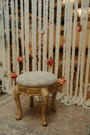 Ornate gold stool with gray upholstery against a backdrop of hanging string lights and roses.