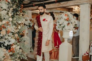 A groom in traditional South Asian attire stands elegantly in a rustic venue with floral decorations.
