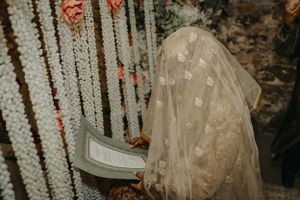 A bride in traditional dress seen through hanging floral decorations and beaded curtains.
