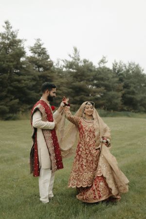 Couple in traditional South Asian wedding attire dances together on grassy field with pine trees.