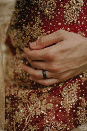 A hand wearing a black ring rests against richly embroidered red and gold traditional wedding attire.