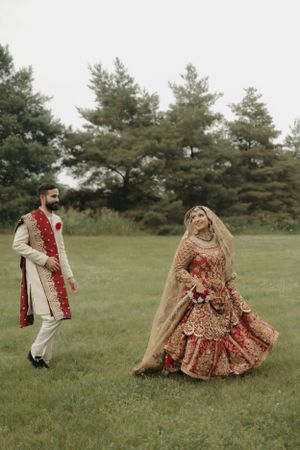 Couple in traditional South Asian wedding attire poses in a scenic outdoor setting with pine trees.