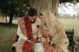 A couple in traditional South Asian wedding attire sits together on a bench in a garden setting.