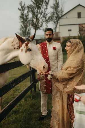 Wedding couple in traditional attire poses with a white horse at a rustic farm setting.