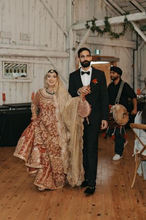 A couple in traditional South Asian wedding attire walks across a rustic wooden floor.