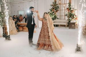 Couple dancing at reception in traditional South Asian wedding attire with white backdrop and floral decor.