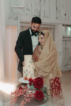 A couple in traditional wedding attire stands beside an elegant white cake decorated with roses.