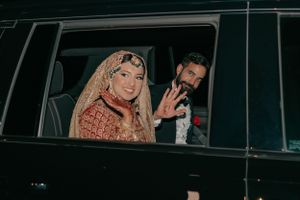 A couple in wedding attire waving from inside a black vehicle during their departure.