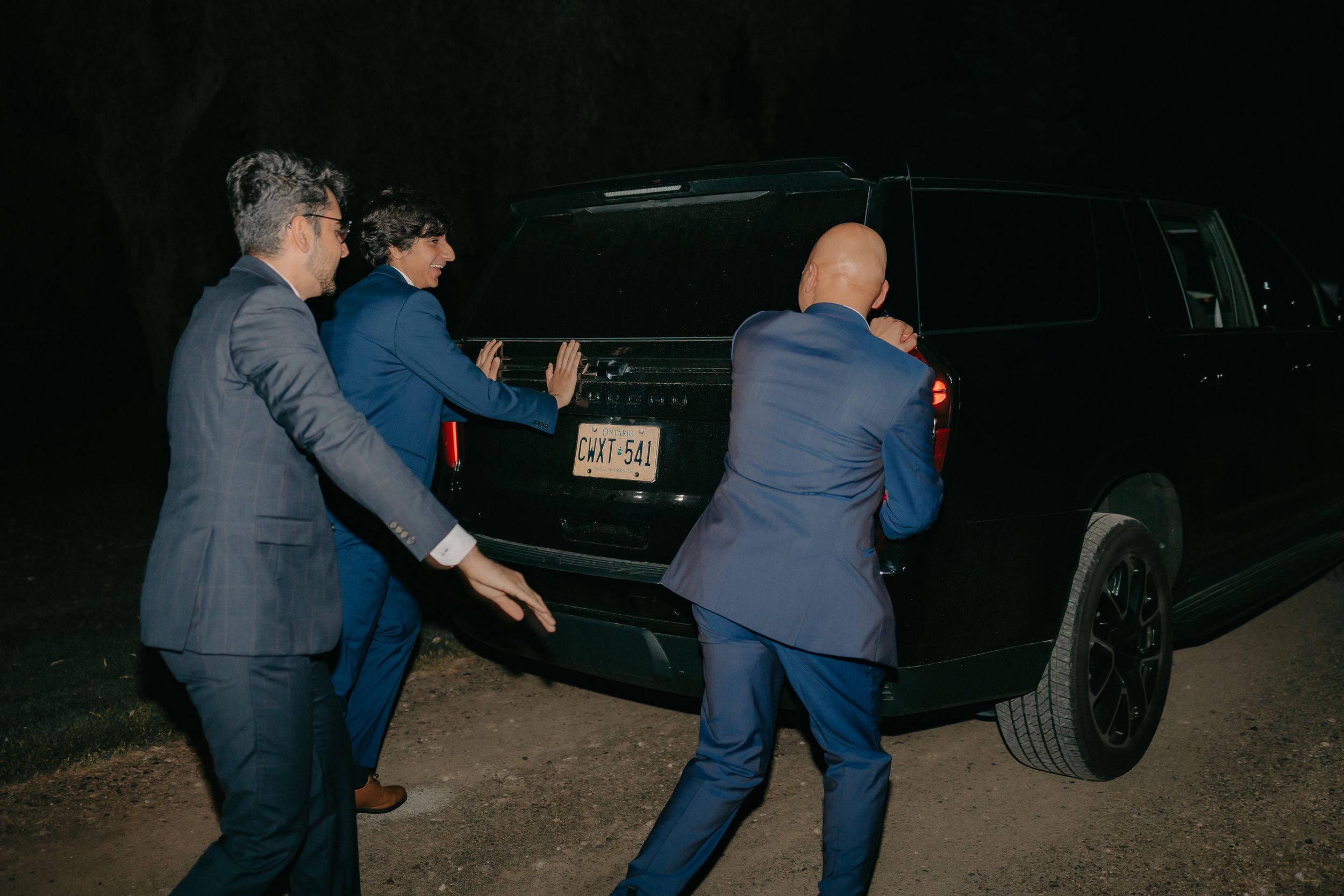 Three people in formal attire pushing a car in a nighttime scene.