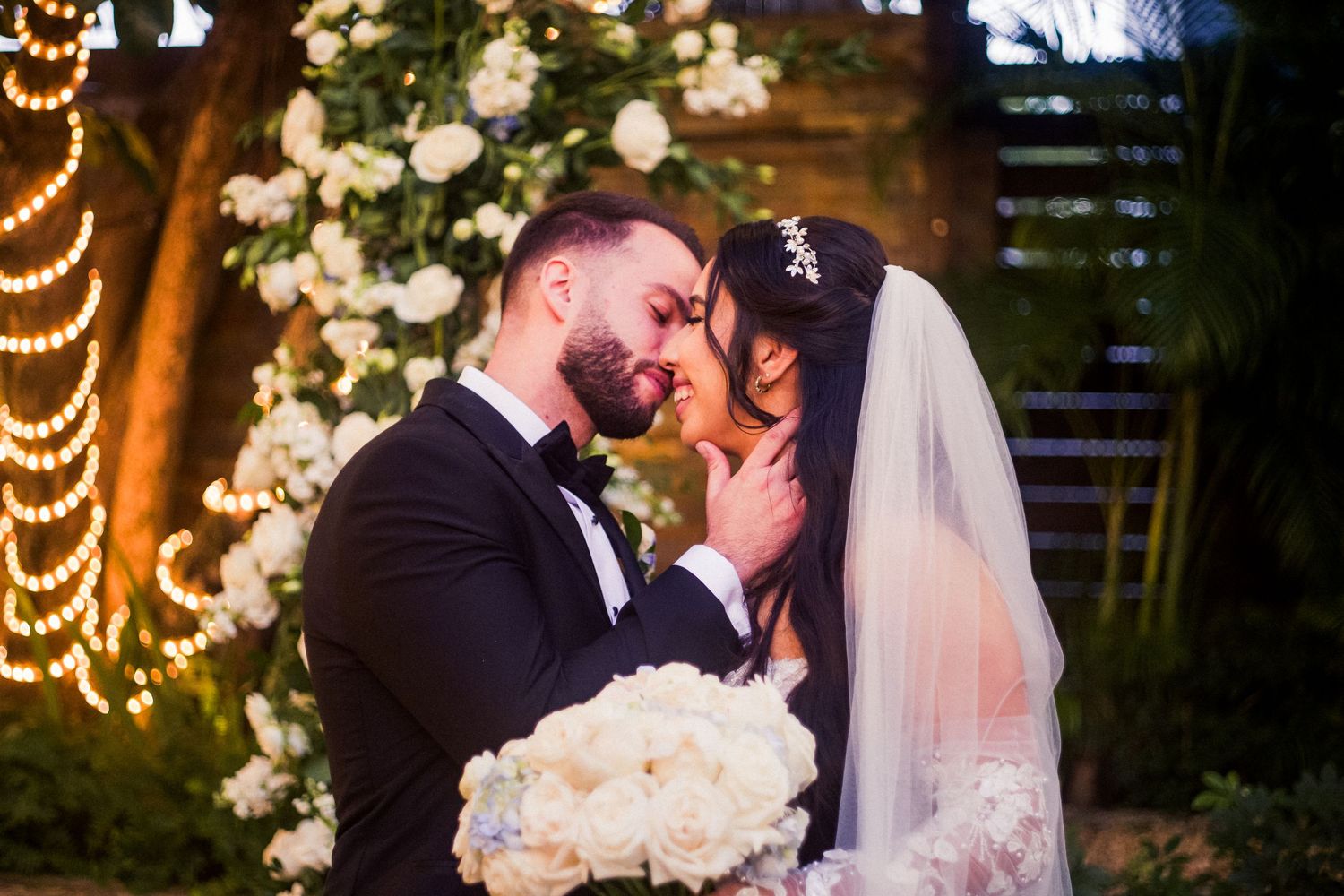 A romantic wedding moment captured with string lights and white floral decor in an outdoor garden setting.