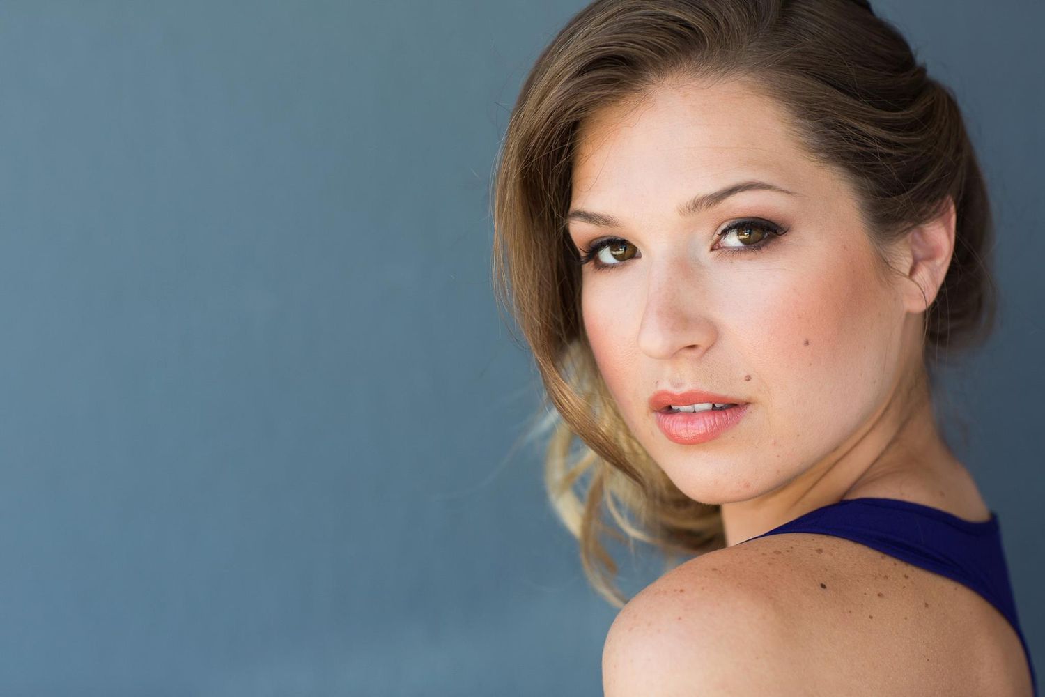 A person in a navy blue dress poses against a light colored background with professional makeup and styled hair.