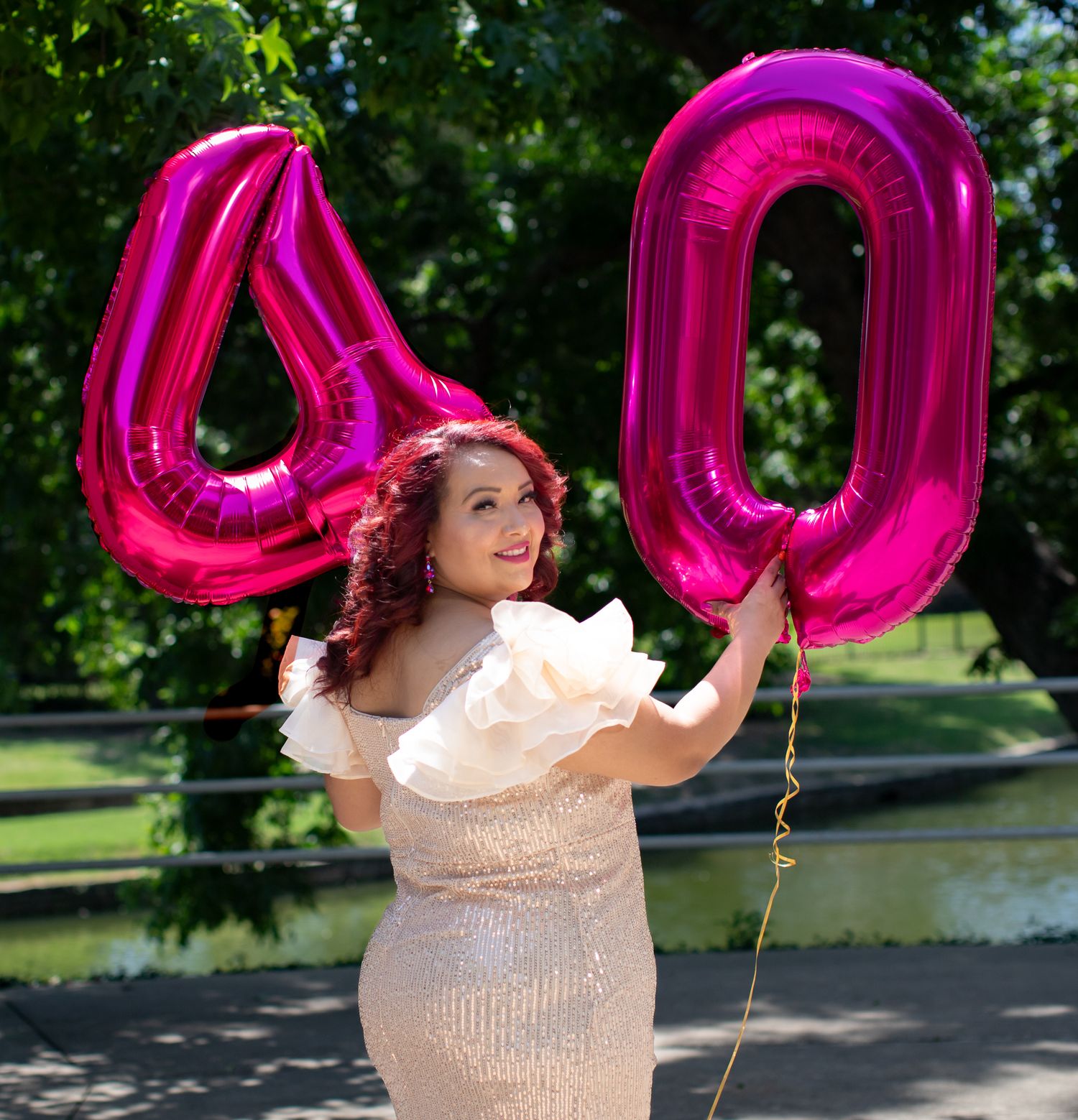 Large pink 40th birthday number balloons held by someone in a sparkly cream-colored dress against a sunny outdoor backdrop.
