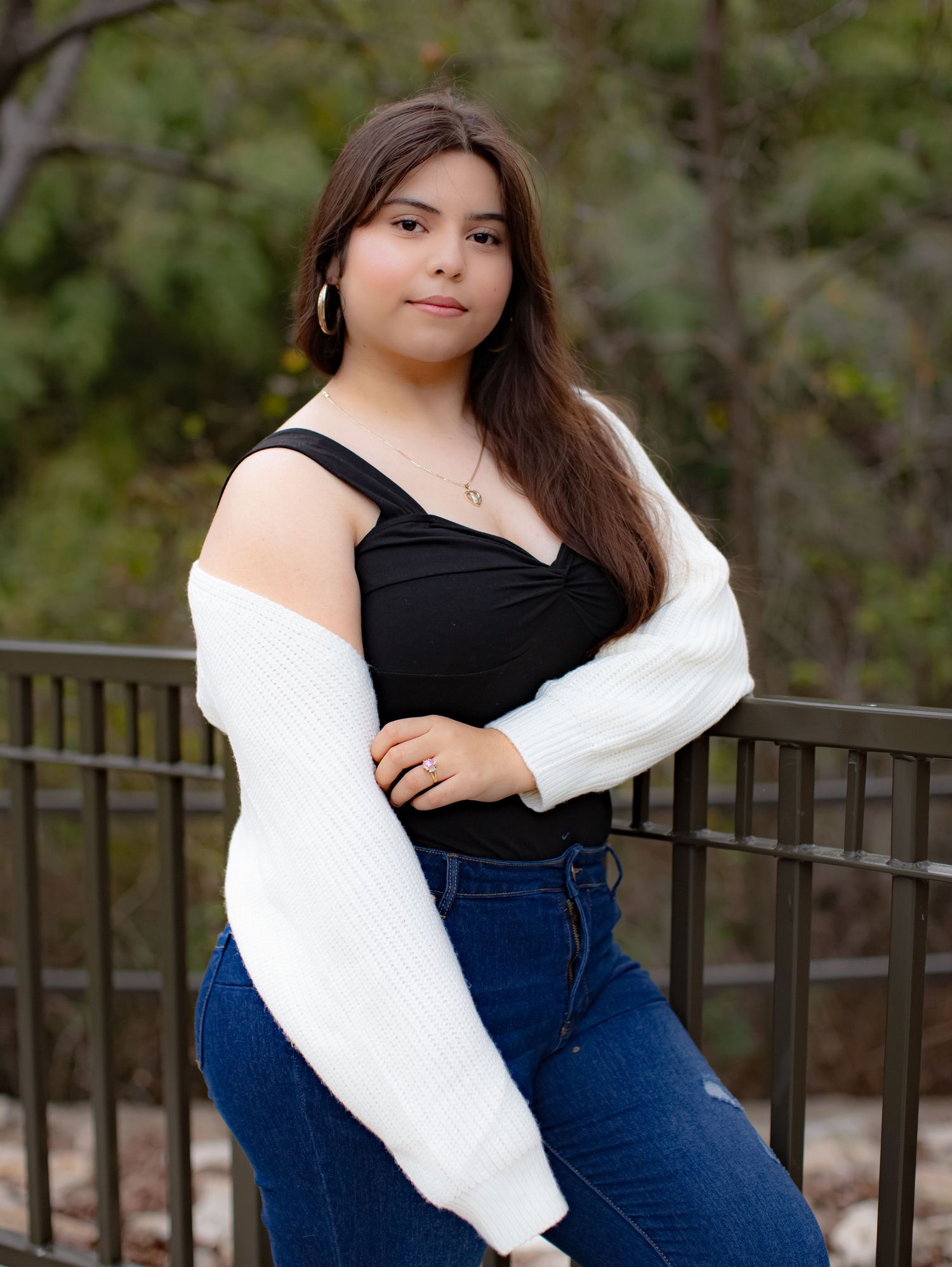 Young person in a black sleeveless top and white cardigan poses outdoors against an iron railing in natural lighting.