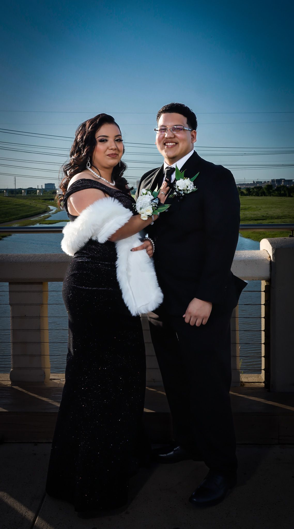 Two people in formal black attire pose together on a bridge at dusk wearing boutonnieres and a white fur wrap.