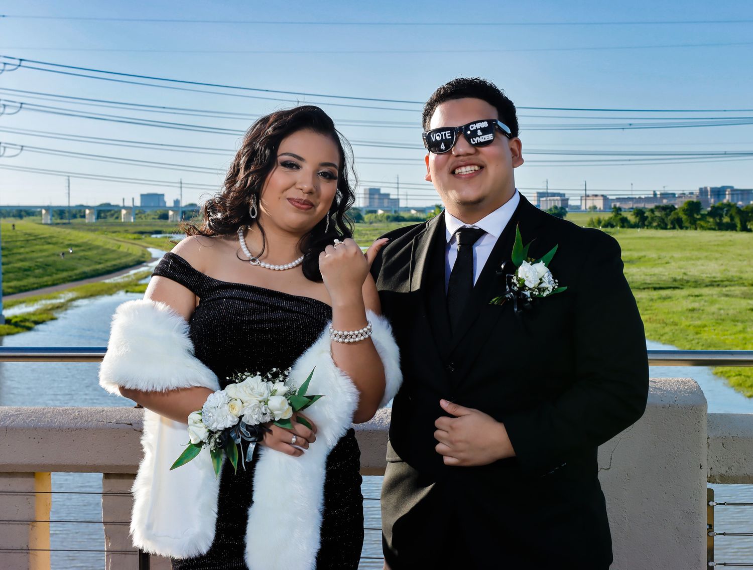 A formal couple poses outdoors in wedding attire, with white flowers and power lines visible in the background.