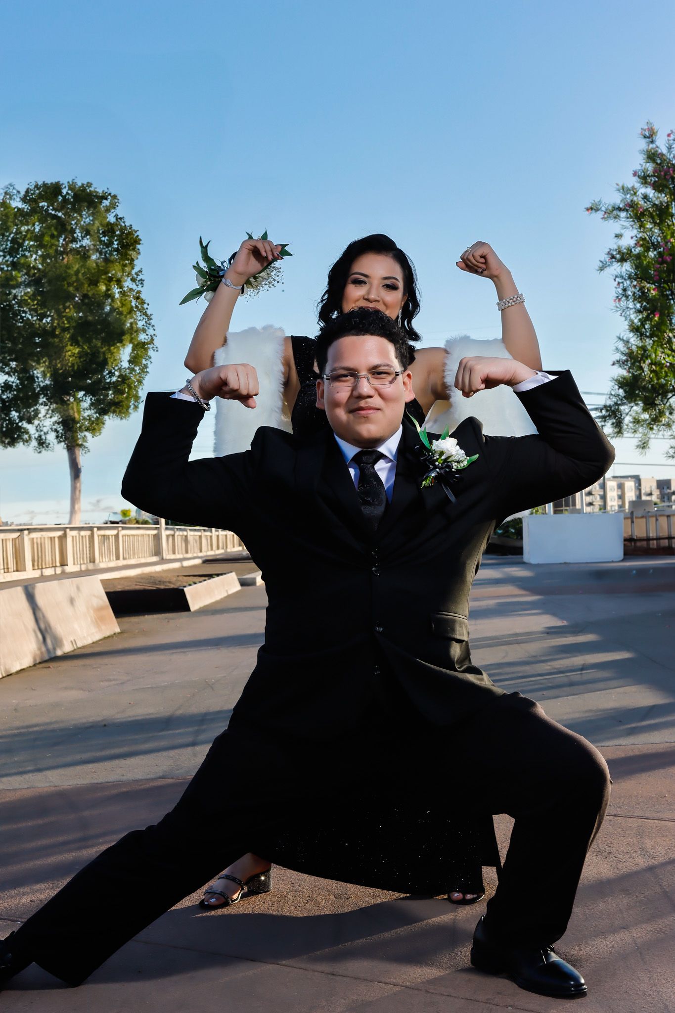 A playful couple poses together outdoors, both flexing their arms in a fun celebratory stance on a sunny day.