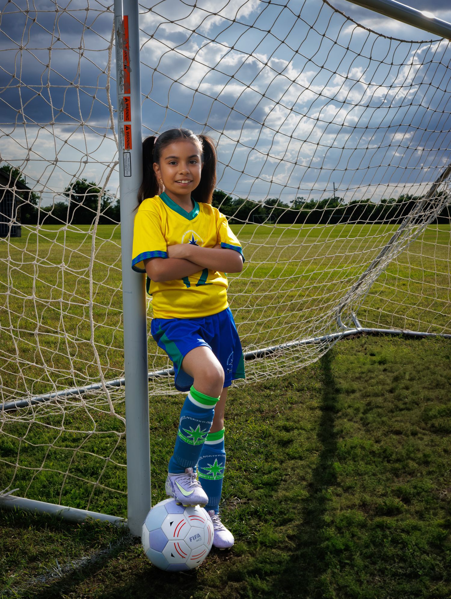 Soccer player in yellow and blue uniform poses with ball at goal post on sunny grass field.