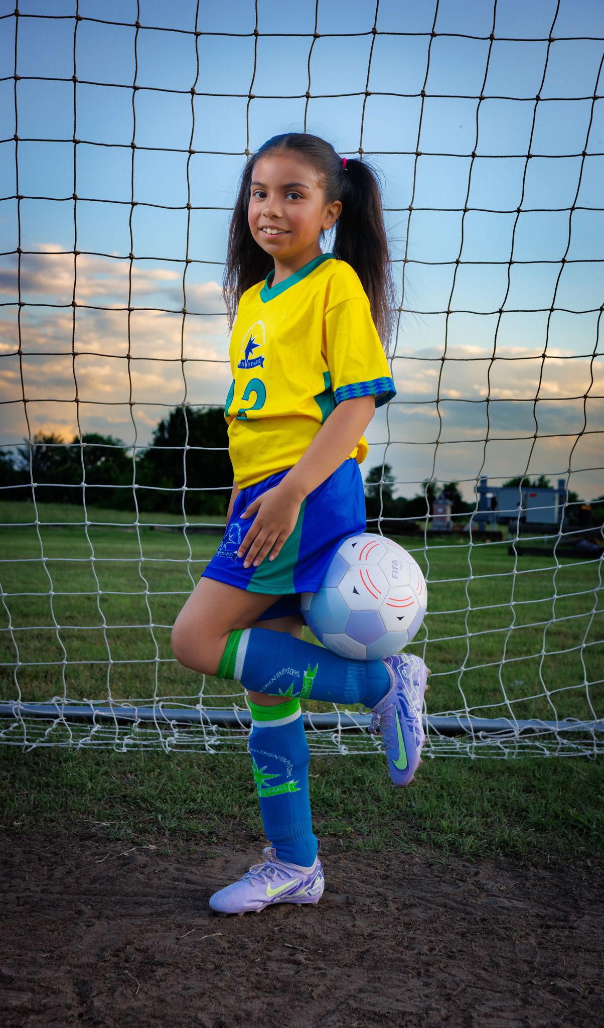Young soccer player in yellow and blue uniform poses with ball against goal net during sunset practice session.