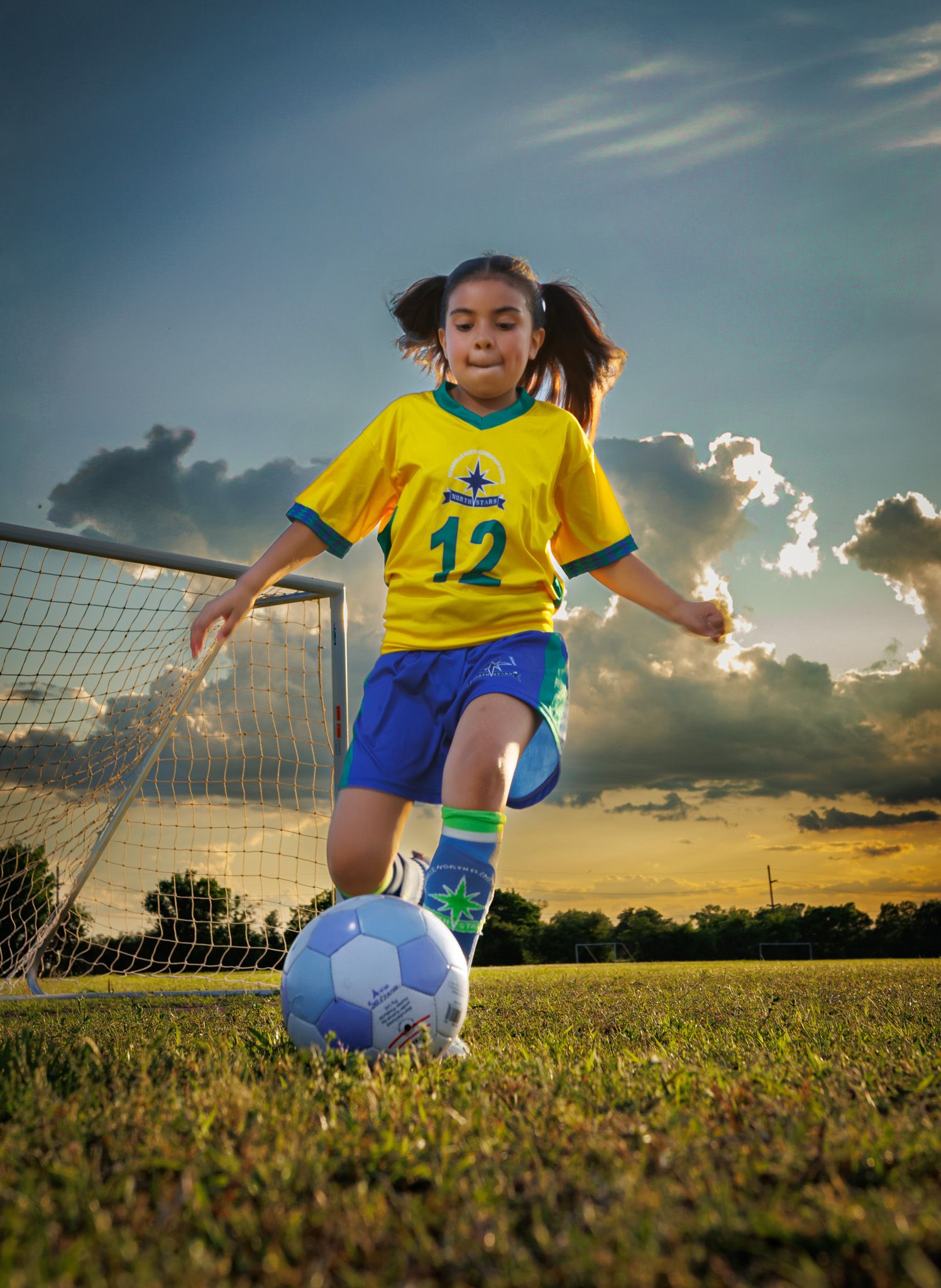 Soccer player in yellow jersey number 12 and blue shorts kicks ball during dramatic sunset with goal net in background.