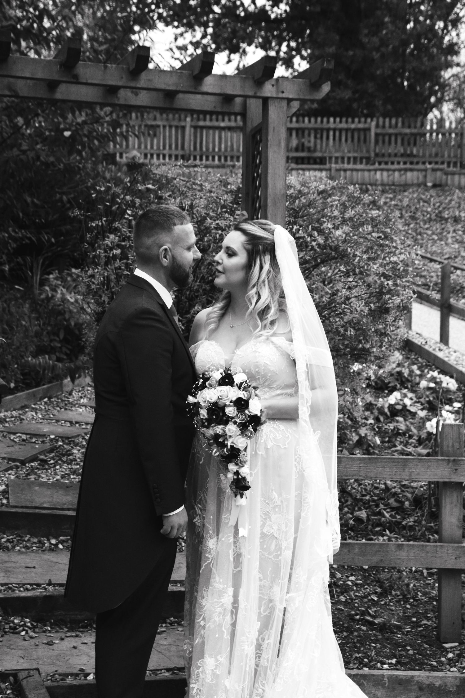 A romantic black and white wedding portrait of a couple embracing outdoors beside a wooden fence.