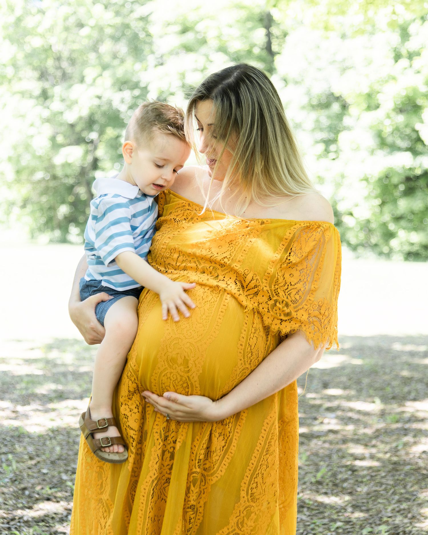 A pregnant woman in a yellow lace dress holds a small child wearing blue and white stripes in a sunlit outdoor setting.