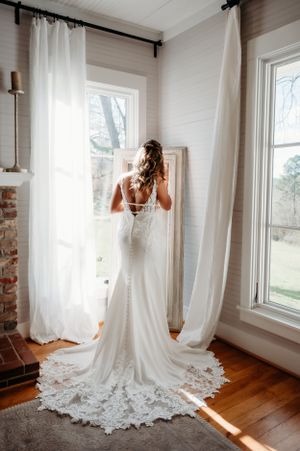A bride in a fitted lace gown stands by windows, showing the detailed back of her dress.