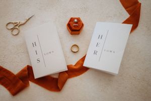 Wedding vow books and rings styled with rust-colored ribbon and gold scissors on cream background.