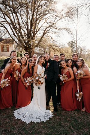 Wedding party poses together outside with bridesmaids in burgundy dresses and groomsmen in dark suits.