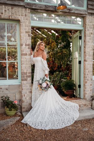 A bride in a lace off-shoulder wedding dress poses in front of a rustic brick greenhouse entrance.