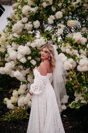 A bride in a lace dress poses against a backdrop of blooming white hydrangea bushes.