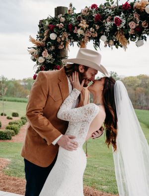 A couple shares a romantic kiss under a floral arch in an outdoor wedding setting.