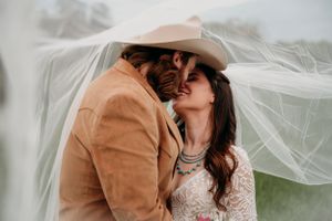 A couple embraces under flowing wedding veil with groom wearing brown blazer and cowboy hat.