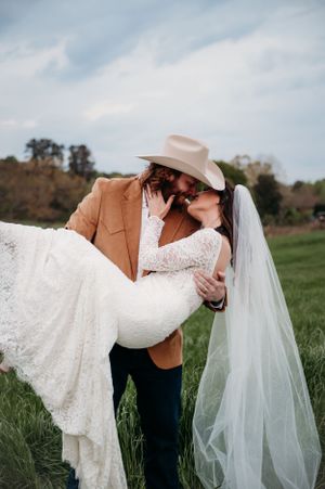 A groom in tan jacket lifts his bride in a romantic pose on a grassy hillside.