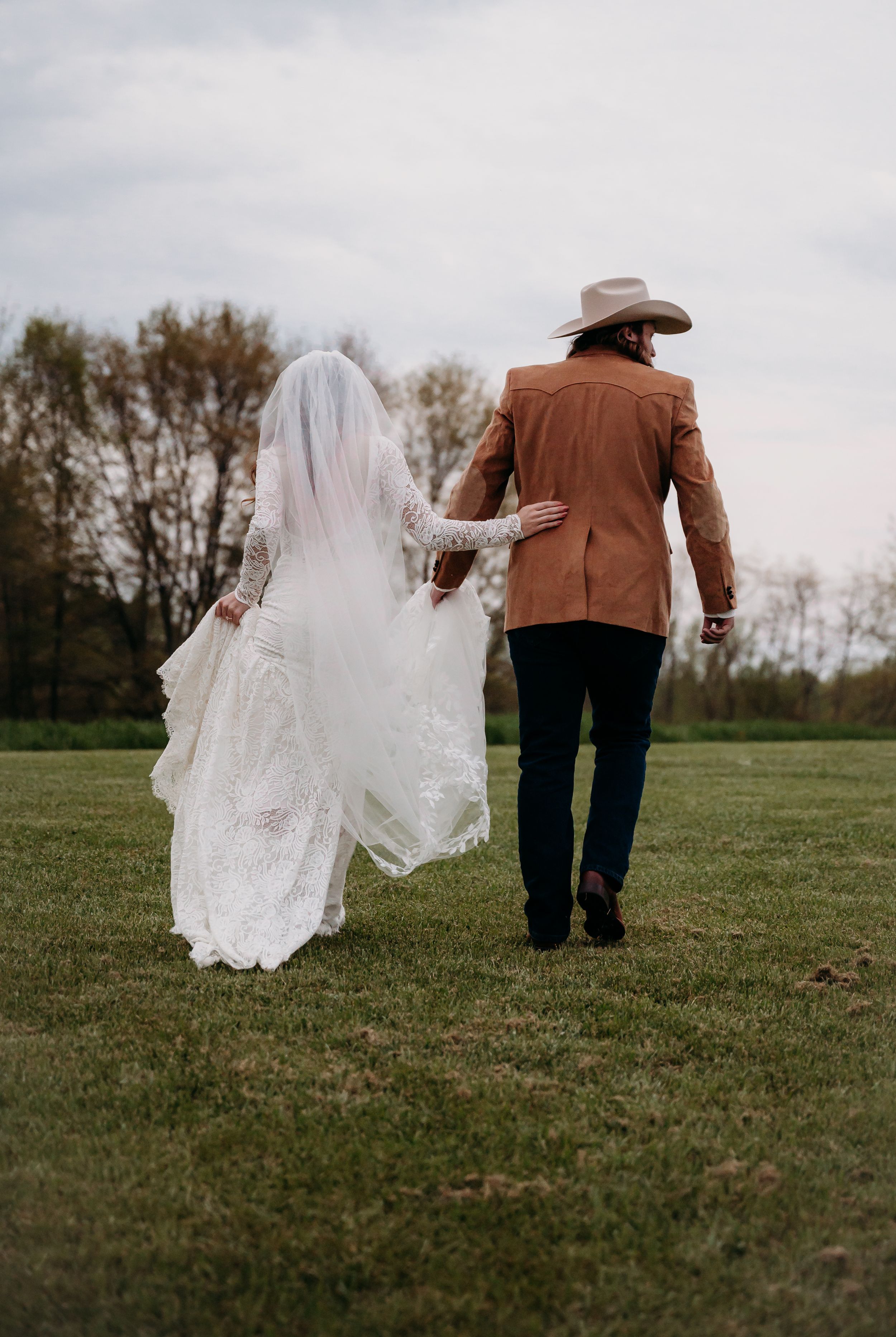 A newlywed couple walks hand in hand across a grass field with flowing white veil trailing behind.