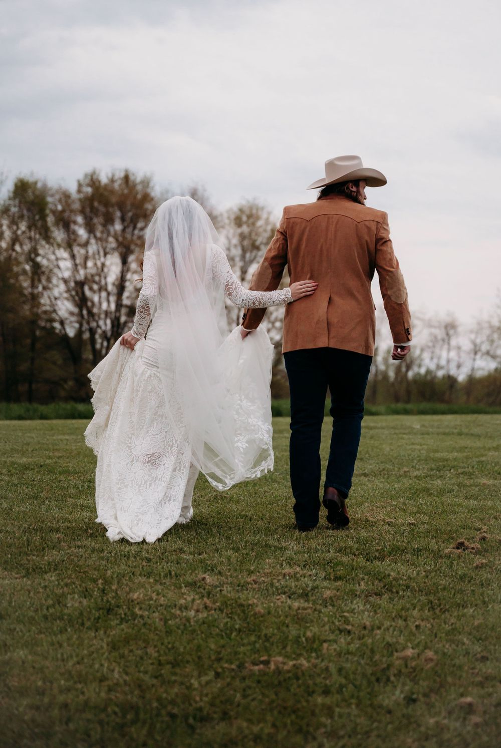 A newlywed couple walks hand in hand across a grass field with flowing white veil trailing behind.