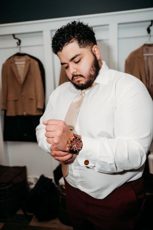 A person in a white dress shirt adjusts their cuff links while standing in front of hanging jackets.