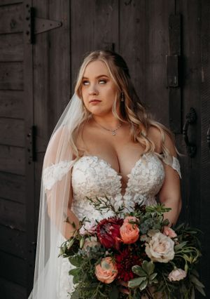 A bride in a lace wedding dress holds a vibrant bouquet of roses against a rustic barn backdrop.
