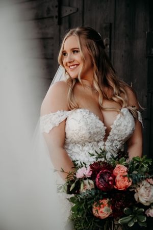A bride poses with a colorful bouquet featuring burgundy and peach roses against rustic wooden walls.
