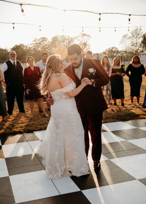 A couple shares their first dance on a checkered dance floor at sunset with string lights overhead.