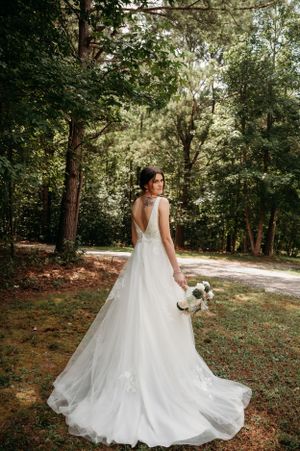 Bride in flowing white gown poses in a sunlit forest setting holding bouquet.