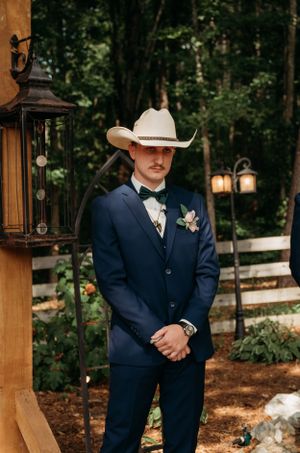 Groom in navy suit and white cowboy hat stands next to rustic lantern decor.