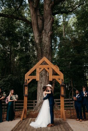 Wedding ceremony under wooden arch with couple sharing first kiss surrounded by wedding party.