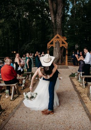 Wedding couple shares romantic dip kiss under rustic wooden arch as guests applaud.