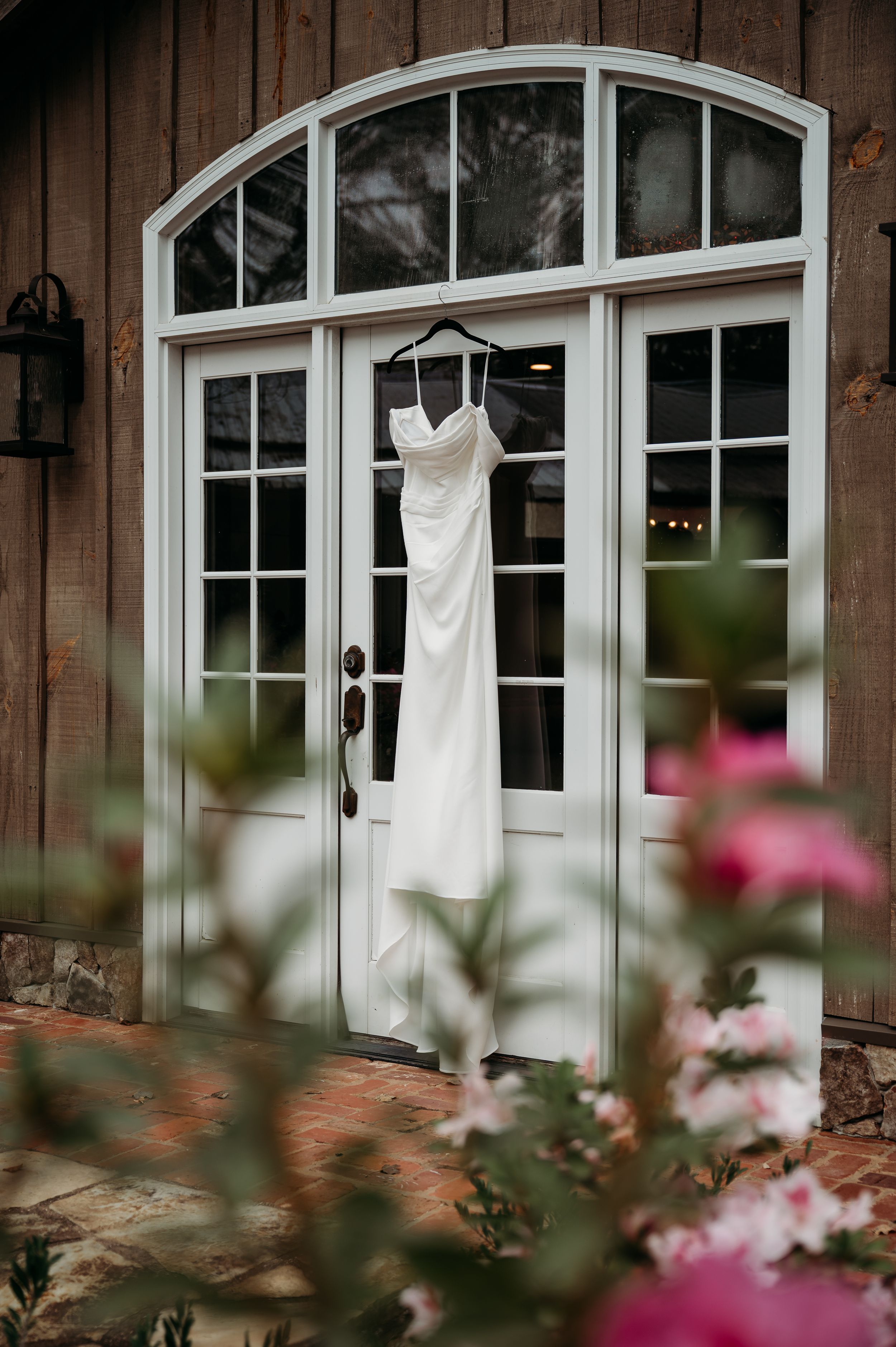 A white wedding dress hangs on an elegant arched doorway with flowers in the foreground.