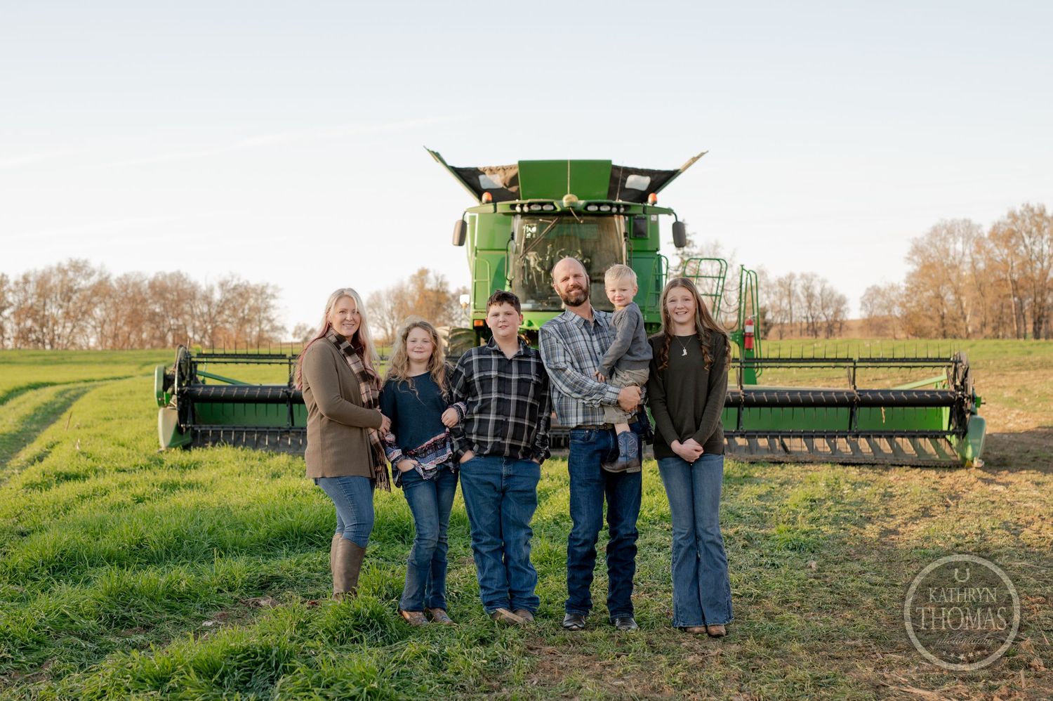 A family stands together in front of a large green combine harvester in a field.