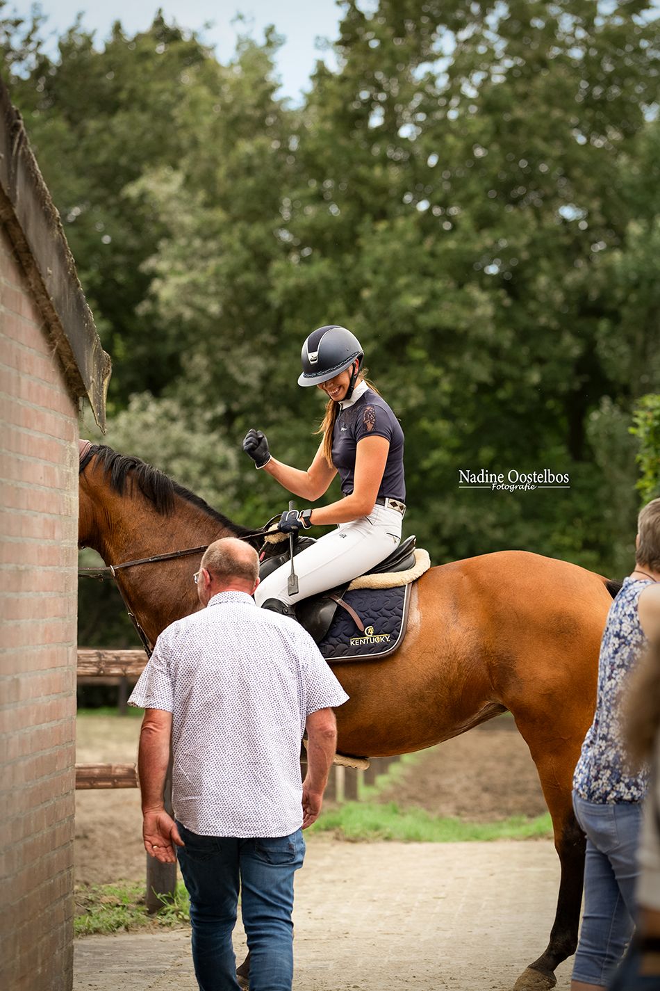 Rider mounted on chestnut horse while consulting with others near stable wall.