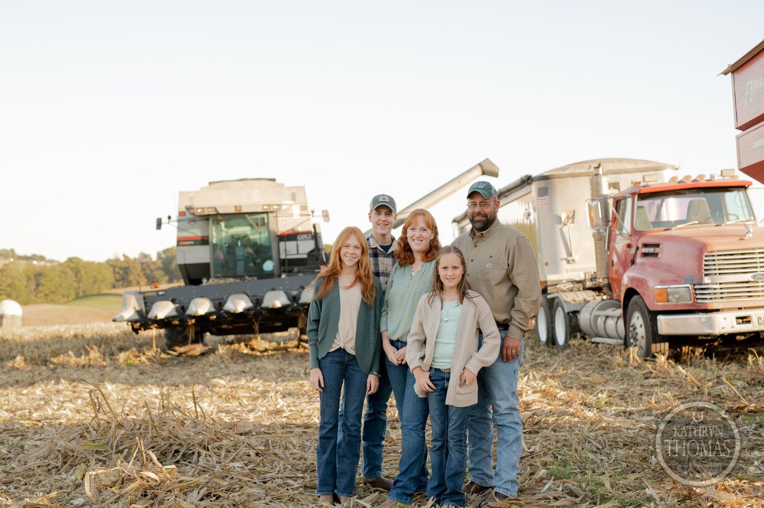A family group poses together in front of farm equipment and grain storage facilities at sunset.