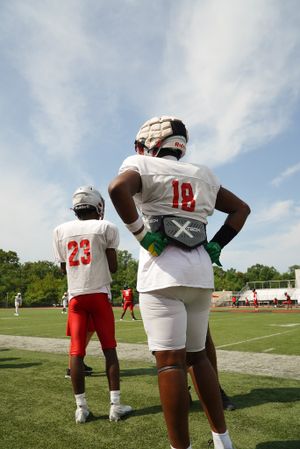 Two football players in white uniforms stand on the sideline during practice.